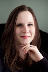A picture of a white woman with straight brown hair and brown eyes. She has her hand placed under her chin and is smiling softly in a black chair in front of a gray wall. There is a slightly sepia filter over the photo.