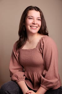 Photo of a smiling white woman with long brown hair, wearing a long sleeve pink shirt.