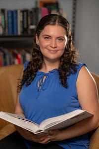 A woman with curly brown hair sits in an armchair in front of a bookshelf. She is holding an open book in her lap.