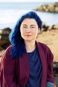 Portia, a white woman with a round face and curly blue hair, sits in front of a curve of rocks and ocean. She wears a burgundy jacket, a blue top, and long silver earrings. The wind has blown her hair over one shoulder and she looks directly at the camera.
