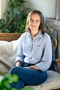 The photo shows a smiling white woman with shoulder-length brown hair sitting on a sofa with a plant in the background. She is wearing a light blue shirt.
