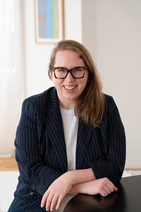 Anna McCallie, a white woman with dark blonde hair, sits in a brightly lit room with white walls. She is wearing a navy blue pinstriped blazer and has her arms crossed over one another as they rest on a table. She wears large tortoiseshell glasses and is smiling at the viewer.