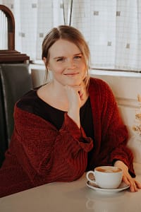A white woman in her early 30s with dark blonde hair and blue eyes. She is sitting at a booth with a cup of coffee, wearing a dark purple top and a brown cardigan.