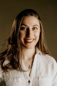 A photo of Deena smiling. She has brown hair and wears a white button-up shirt in front of a dark brown background.