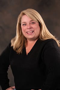 A seated, smiling woman with blonde hair, blue eyes and white skin, wearing a v-neck black sweater.