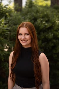A woman in a black top and long red hair standing outside in front of green plants.