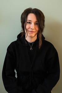 T. E. Forest, a white person with dark brown hair, wearing a black sweater and a silver moon necklaces looks wryly into the camera.
