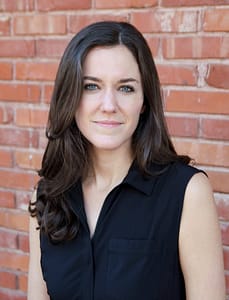 A woman with long, wavy brown hair and blue eyes stands in front of a red brick wall. She is wearing a sleeveless black blouse with a zippered front and a collar. She has a neutral expression and is looking directly at the camera.