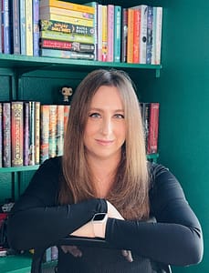 Author Michelle C. Harris poses in front of a dark green bookshelf filled with an assortment of colorful books. She has straight, dark strawberry blond hair and smiles softly at the camera with her arms crossed over the top of a desk chair.