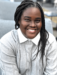 A dark-skinned Black woman with her hair in black box braids that go down to her mid-back, falling over her left shoulder. She is smiling at the camera while wearing a white collared gray sweater dress, in front of a blurred background.