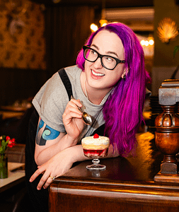 Helen Gaskell, a white woman with long bright purple hair, stands at a bar eating a miniature trifle. She is looking over her shoulder, laughing at an unseen figure, probably her ludicrous husband.