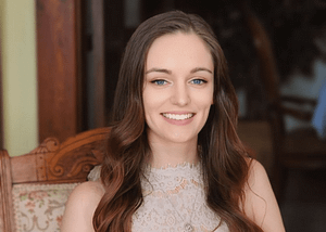 Photo of a white woman with long, brown hair and blue eyes. She is shown smiling in a pink dress while sitting in an antique chair.