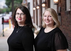 A picture of two white women in their mid-thirties, standing outside next to local businesses. The one on the left has shoulder-length brown hair, glasses, and blue eyes. She is shown from the chest up, smiling, and is wearing a black long-sleeve shirt. The one on the right has just above the shoulder blonde hair and blue eyes. She is shown from the chest up, smiling, and is wearing a short-sleeve black shirt.