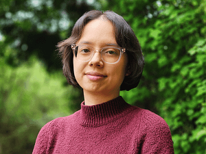 A photo of a half Chinese, half white woman with short, dark-brown hair, brown eyes with clear frame glasses, and a maroon turtleneck sweatshirt. She is shown from the chest up and is standing in front of some green trees in the woods.