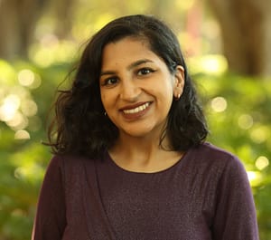 A photo of a South Asian woman with shoulder-length, wavy black hair and brown eyes in her late twenties amongst greenery. She is shown from the chest up, smiling, and wears a purple, long-sleeved top.