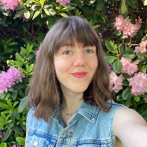 A photo of a white, freckled woman smiling into the camera. She has brown, shoulder-length hair, bangs, blue eyes and is standing in front of a pink flower hedge.