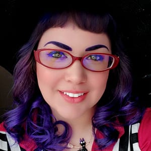 Close up photo of a smiling woman with red glasses, curled purple hair, and green eyes wearing a witch hat, red blouse, and a black cat necklace.